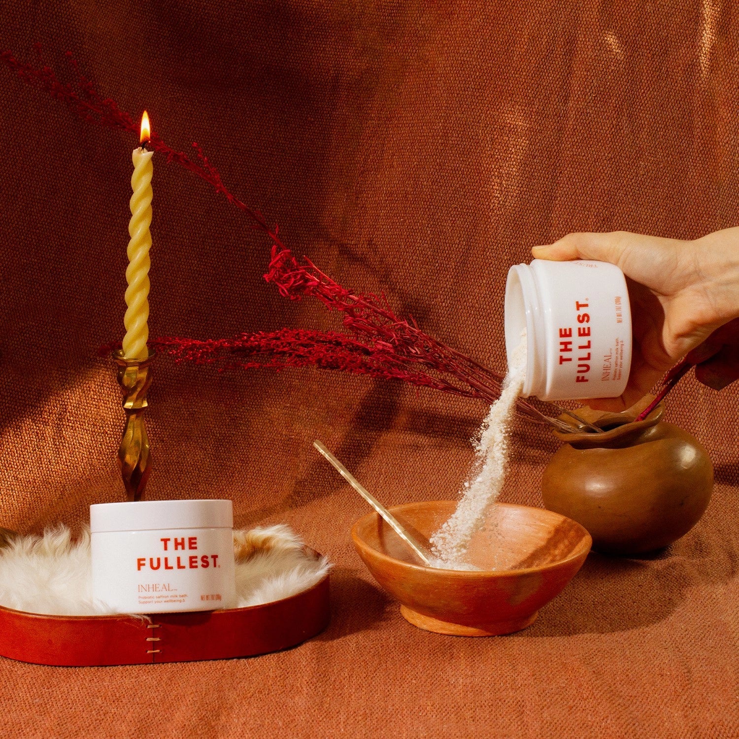 A hand pours Inheal Milk Bath from a container labeled "THE FULLEST" into a ceramic bowl on a fabric surface. Nearby, another container and a twisted candle in a holder are placed beside dried red flowers. The setup exudes a warm, rustic ambiance perfect for indulging in skin hydration.