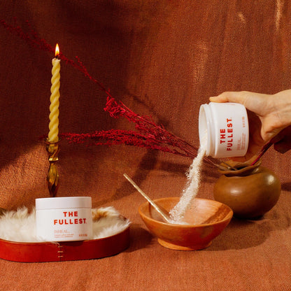 A hand is pouring Inheal® Probiotic Saffron Milk Bath into a wooden bowl from a container labeled "THE FULLEST." A lit twisted candle and red dried flowers are placed to the left, with a textile backdrop of warm, rust-colored tones.