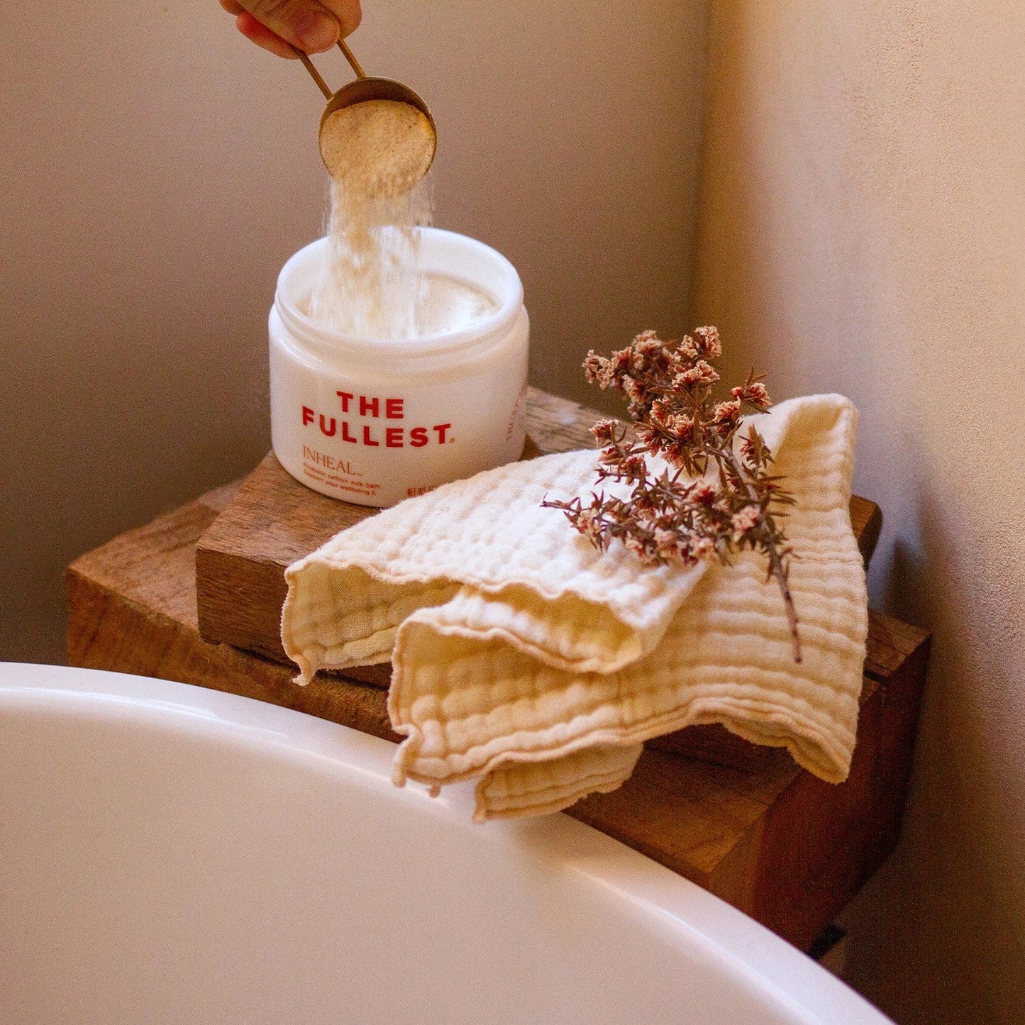 A hand pours THE FULLEST's Inheal Milk Bath, enriched with probiotics and saffron, from a wooden scoop into a white jar labeled "Inheal." The jar rests on a wooden plank beside a white bathtub. Two beige textured cloths and a sprig of dried flowers are also placed on the plank, enhancing the scene's focus on skin hydration.