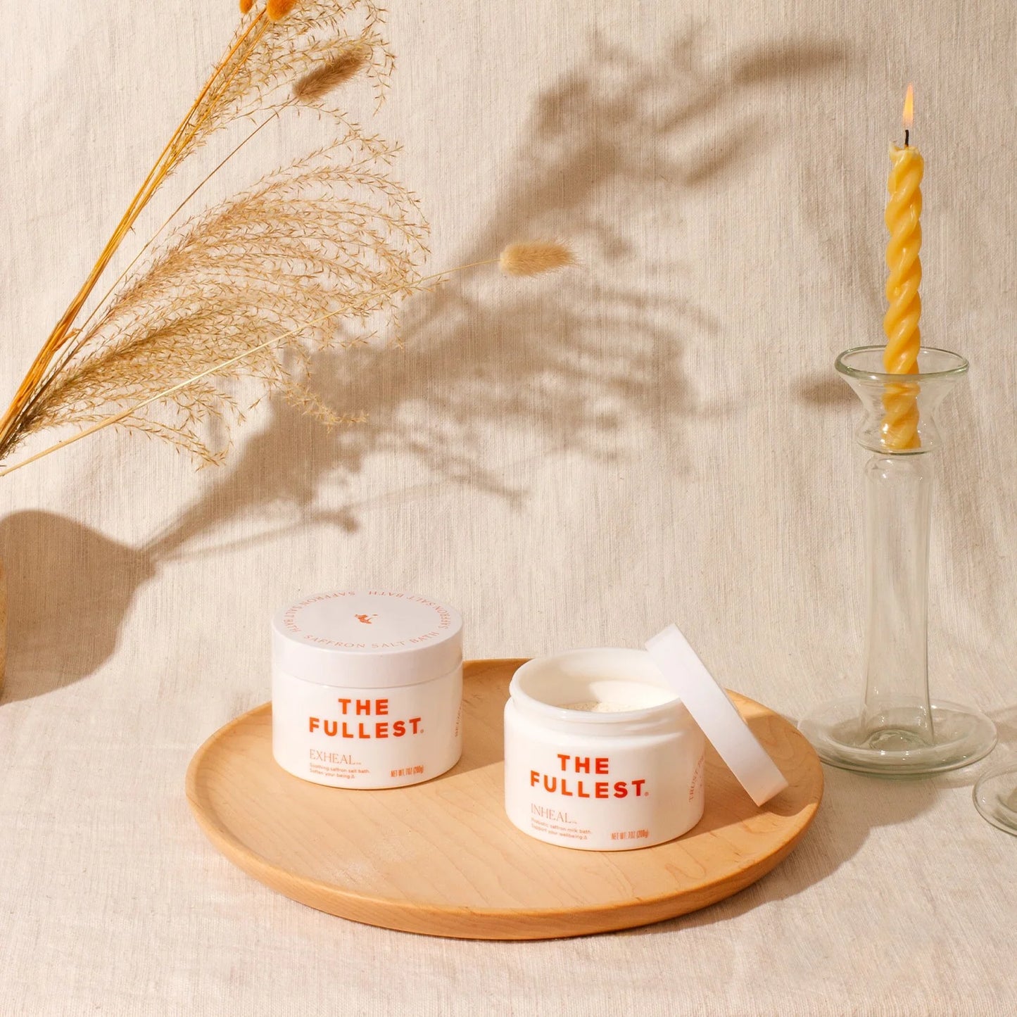 A wooden plate holds two jars of Inheal® Probiotic Saffron Milk Bath by THE FULLEST, celebrated for their nourishing benefits. One jar is open, showcasing the rich cream within. To the right stands a tall, twisted candle in a clear holder, while dried pampas grass adorns the left, all arranged on a beige fabric backdrop.