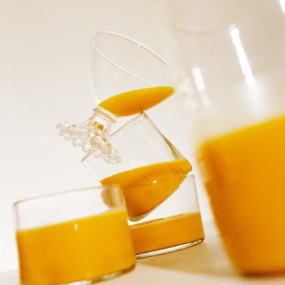 A titled wine glass containing THE FULLEST's Warm Feelings Bulk saffron latte is precariously balanced on a horizontal glass, also filled halfway with the same saffron latte. In the foreground, there's a blurred close-up of another glass filled with Warm Feelings Bulk saffron latte on the right side.