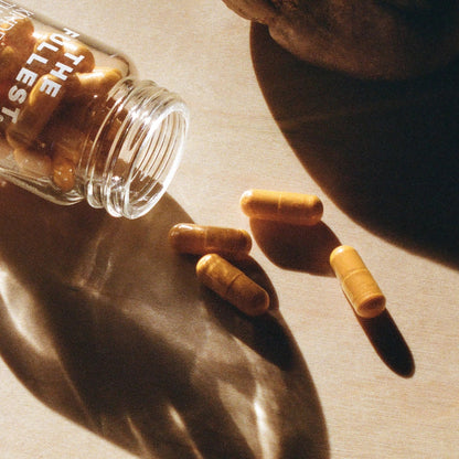 A glass bottle with capsules lies on its side, spilling some saffron supplement onto a surface. The THE FULLEST™ Kinder Thoughts label is partially visible, and the scene is illuminated by warm, natural light casting shadows.