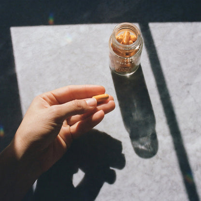 A hand holds an orange capsule above a marble surface bathed in sunlight. To the right, an open glass bottle of THE FULLEST's Kinder Thoughts saffron supplement is positioned, casting a shadow on the surface. The shadows of both the bottle and the hand are visible.