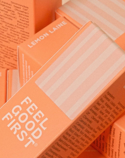 A close-up of the product packaging showcases the vibrant orange and white design with "LEMON LAINE" and "FEEL GOOD FIRST" prominently displayed. The packages, containing the luxurious Botanical Bounce Glow Moisture Face Oil Serum, are stacked and arranged at different angles. Partial text detailing product information is visible on the sides.