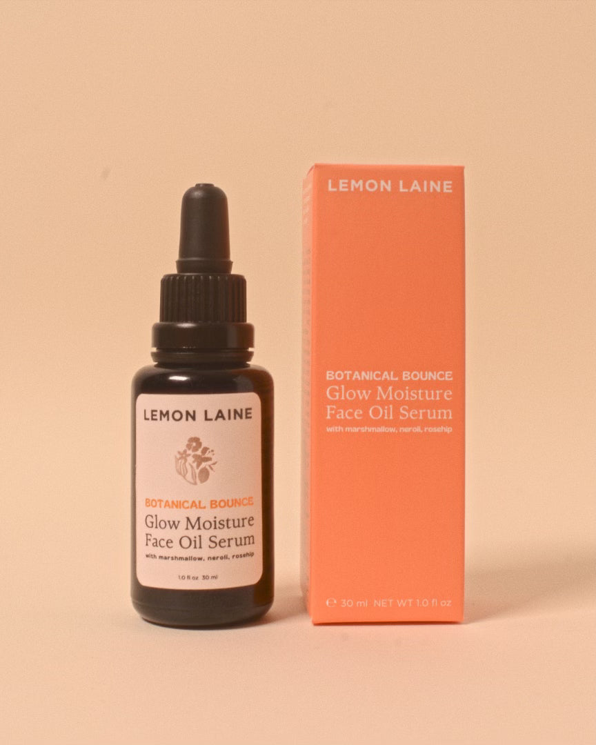 A bottle of LEMON LAINE Botanical Bounce Glow Moisture Face Oil Serum is placed next to its orange box. The serum bottle, featuring a black dropper top and a white label with brand and product information, highlights the luxe botanicals used in this vegan skincare gem. The box mirrors the branding and text of the label.