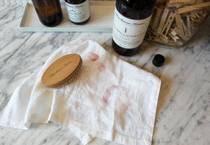 A white cloth with lipstick stains lies on a marble surface, a Murchison-Hume Compostable Laundry Brush with natural bristles placed on top. Near the cloth, bottles of stain remover and a jar filled with clothespins complete the scene.