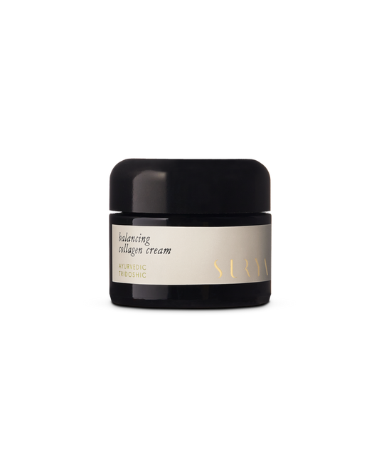 A jar of Surya Balancing Collagen Cream, enriched with Ayurvedic Tridoshic properties and botanical retinol, is displayed against a black background. The natural face cream's black jar features a light-colored label adorned with gold text.