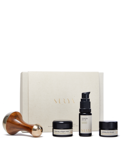 The image showcases the Surya Skincare Discovery Set, featuring a wooden-handled Kansa Wand, a jar labeled "balancing collagen cream," a bottle labeled "balancing face serum," and a jar labeled "nourishing lip therapy," all elegantly arranged in front of a beige Surya box.