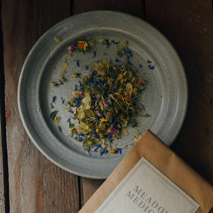 A grey plate holding a colorful mix of dried flowers and herbs is placed on a rustic wooden surface. In the bottom right corner, a partially visible brown pouch labeled "MEADOW MEDICINE TEA BLEND" lies beside the plate.