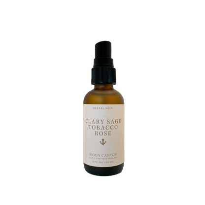 A brown bottle with a black spray nozzle labeled "Clary Sage, Rose & Tobacco Herbal Mist" by Moon Canyon Healing, containing 2 fl oz (60 ml) of liquid in an energetic mist. The bottle is set against a black background.