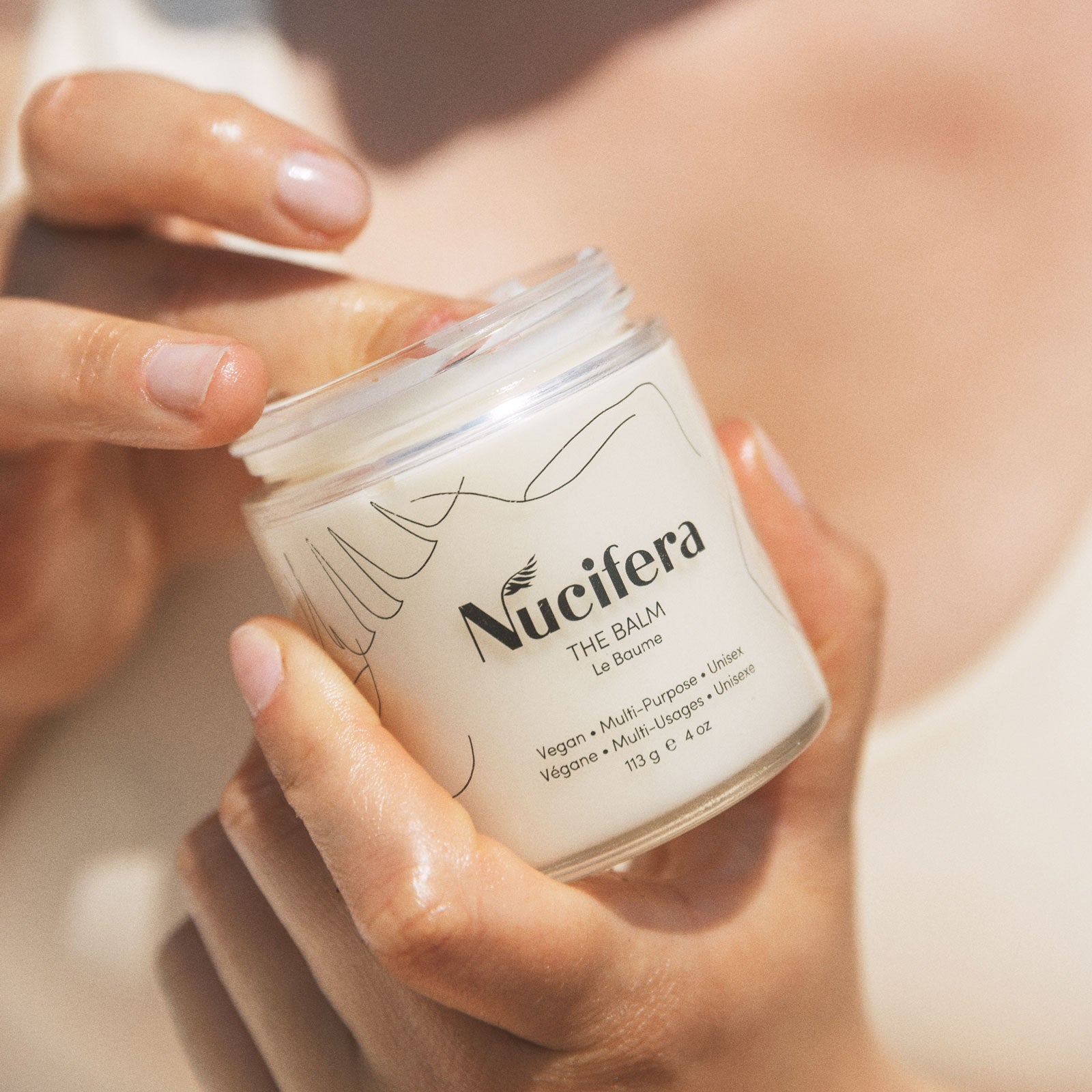 A person is holding and opening a jar of Nucifera Body Store's The Balm with one finger dipped inside. The label indicates that it is a vegan unisex skincare product, suitable for all skin tones and textures. This multi-purpose balm jar contains 113 grams (4 ounces) of nourishing goodness.