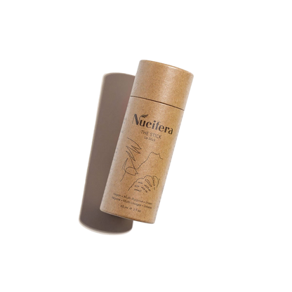 A cylindrical, eco-friendly cardboard container labeled "Nucifera The Stick" from Nucifera Body Store, featuring minimalist line art and holding a solid deodorant stick, is shown against a plain white background. As part of The Essentials Kit, this multi-purpose skincare product appears slightly angled with the lid propped open.