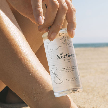 Close-up of a person's hand holding a clear spray bottle labeled "The Mist" by Nucifera Body Store, while sitting on a sandy beach. The bottle, featuring minimalist line art of leaves and faces, delivers a fine mist spray. In the background, the sand and ocean blur into soft focus.