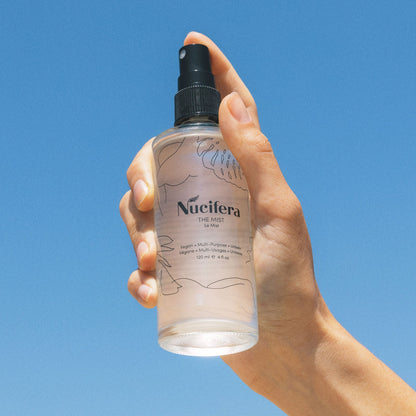 A hand holding a clear spray bottle labeled "Nucifera Body Store The Mist," adorned with minimalist, artistic lines on the label, set against a clear blue sky background. The 100 ml/4 fl oz bottle contains a vegan, multi-purpose fine mist spray that's perfect for refreshing any moment.