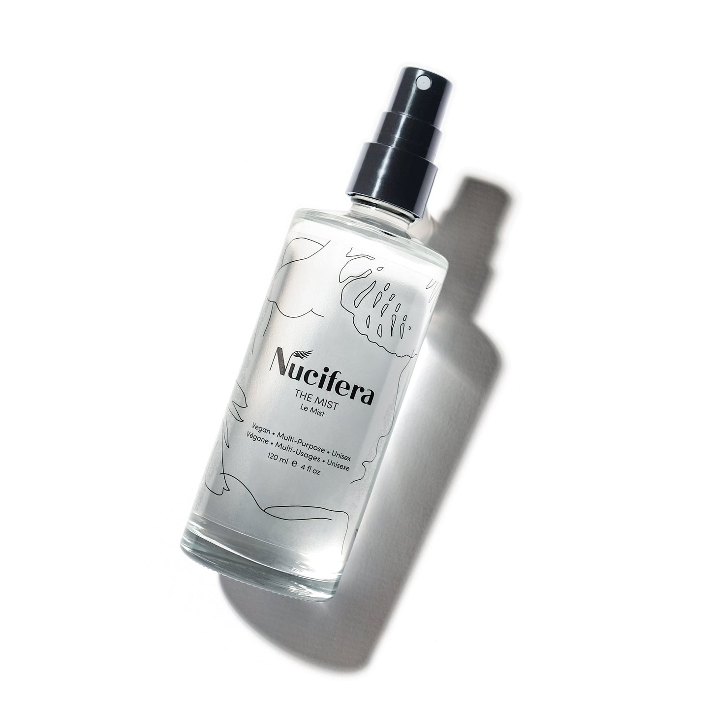 A clear glass spray bottle from Nucifera Body Store, labeled "The Mist," is placed on a white background. The bottle, decorated with intricate botanical line drawings and featuring a black spray nozzle, casts soft shadows that enhance its three-dimensional appearance. The label highlights that this fine mist spray is both vegan and cruelty-free.