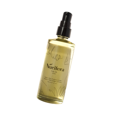 A spray bottle of The Oil from Nucifera Body Store against a black background. This 100ml bottle, part of The Essentials Kit, showcases a clear design adorned with natural, artistic line drawings and boasts a sleek black spray cap. This multi-purpose skincare essential effortlessly integrates into your routine while exuding elegance.