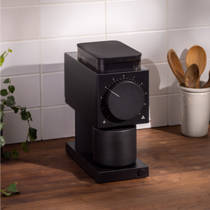 An Ode Brew Grinder by Fellow Wholesale, featuring a sleek black design, Gen 2 Brew Burrs, and 31 grind settings, sits on a wooden countertop against a white tiled backsplash. Nearby, a white ceramic holder contains wooden spoons. A green plant with trailing vines adds a touch of nature to the scene.