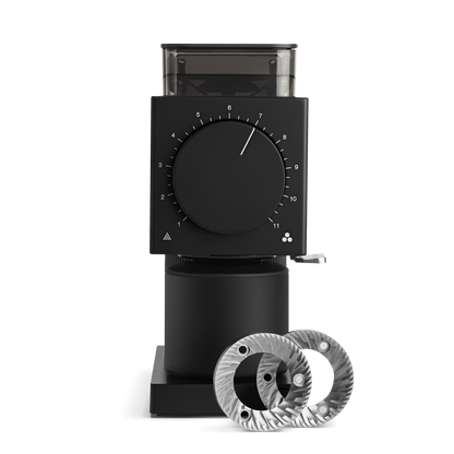 The Ode Brew Grinder by Fellow Wholesale is a sleek, black coffee grinder with a large circular dial on the front set at number 7 and a transparent hopper on top. Featuring Gen 2 Brew Burrs, two shiny, silver burr grinder discs are placed in front of the grinder on a reflective surface.