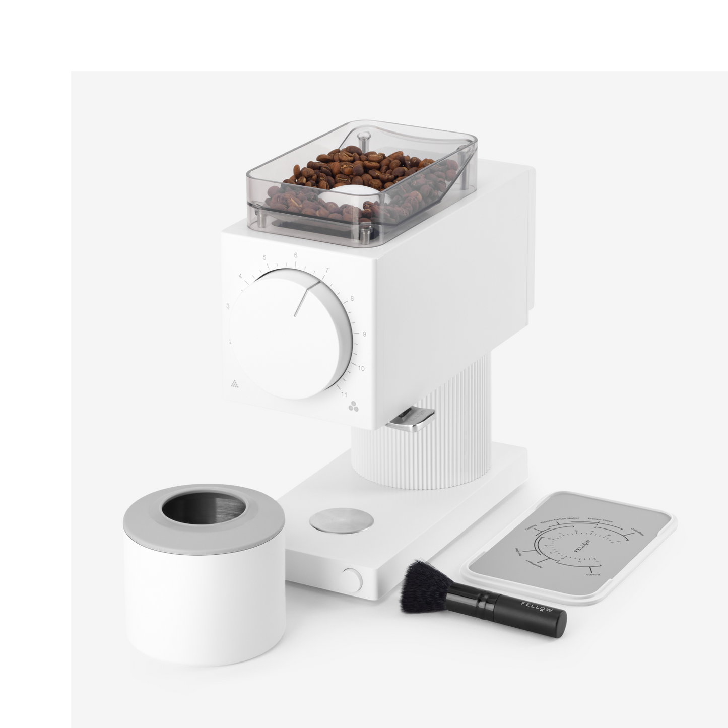 The Ode Brew Grinder by Fellow Wholesale is a modern white coffee grinder featuring a clear container filled with coffee beans on top. It offers 31 grind settings and includes a control knob on the front. Accessories such as a small brush, a removable ground coffee container, and a cleaning tool are also included. Its anti-static technology ensures your countertop stays clean. All items are placed on a white surface.