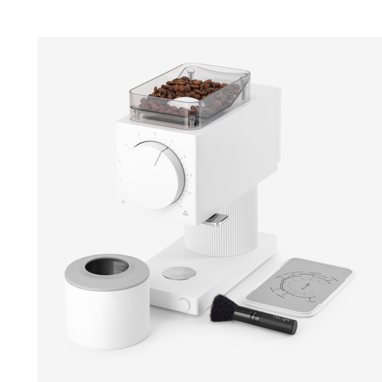 The Ode Brew Grinder by Fellow Wholesale is a modern white coffee grinder featuring a clear container filled with coffee beans on top. It offers 31 grind settings and includes a control knob on the front. Accessories such as a small brush, a removable ground coffee container, and a cleaning tool are also included. Its anti-static technology ensures your countertop stays clean. All items are placed on a white surface.