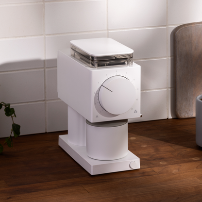 The Ode Brew Grinder by Fellow Wholesale sits on a wooden kitchen counter, boasting a sleek, minimalist design. This modern white coffee grinder features 31 grind settings and anti-static technology, with a clear compartment on top for coffee beans and a large dial for adjusting the grind size. White tiles and a cutting board sit in the background.