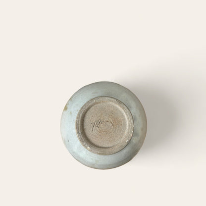 A photograph of the bottom view of a round ceramic vase, showcasing its unglazed beige base with textured concentric circles and a small unreadable mark. The rest of the vase appears to have a light grayish-blue glaze. The background is a simple, off-white surface, reminiscent of vintage goods from Shoppe Maison.