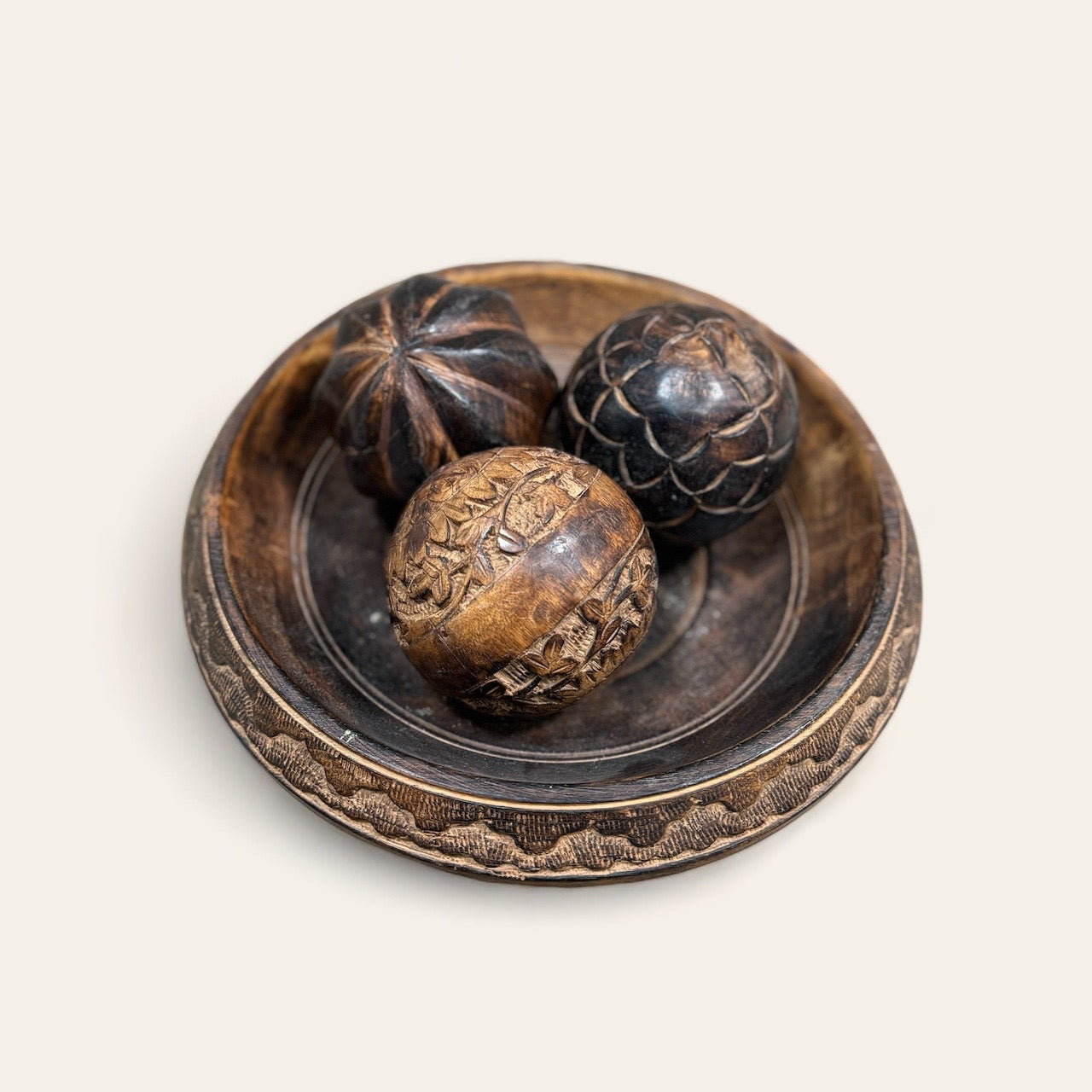 A rustic, dark wooden bowl from Shoppe Maison's Carved Wood Pedestal collection stands gracefully on a vintage wood pedestal, housing three intricately carved balls. Each ball boasts unique patterns and textures that echo the artistry of hand-carved wood. This ensemble embodies a vintage, handcrafted feel against a plain, light background.