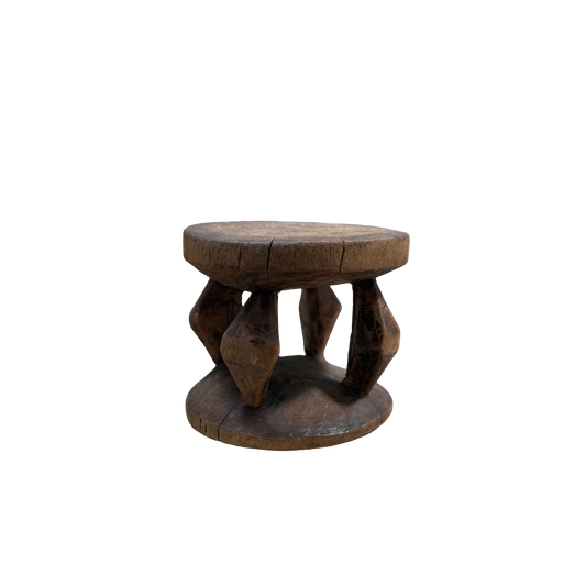 The Vintage African Wood Mini Stool by Shoppe Maison is a small, rustic wooden stool featuring a round top and bottom. It's connected by four intricately carved legs that display geometric shapes in African-inspired designs. The stool's weathered appearance, with visible cracks and wear, enhances its vintage look against the black background.