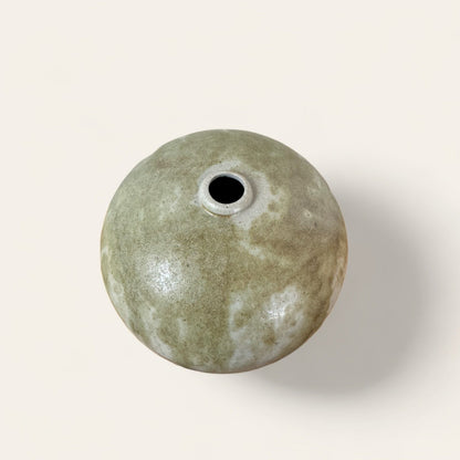 An exquisitely hand-crafted Ceramic Vase by Shoppe Maison, featuring a round shape and greenish hue with a small central opening when viewed from above. The vase boasts a marbled texture that displays an array of varying green and white shades, set against a plain off-white background.