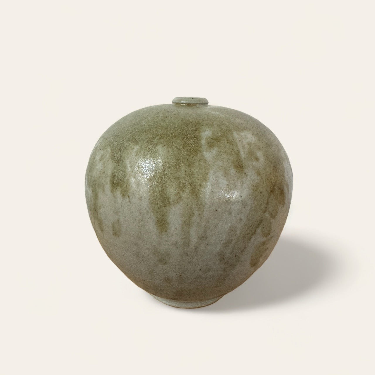 The Ceramic Vase by Shoppe Maison is a hand-made, round ceramic piece with a narrow neck and a small opening. It features a speckled, earthy green and brown pattern that gives it a rustic appearance. The plain, light-colored background accentuates the texture and shape of this vintage-inspired good.