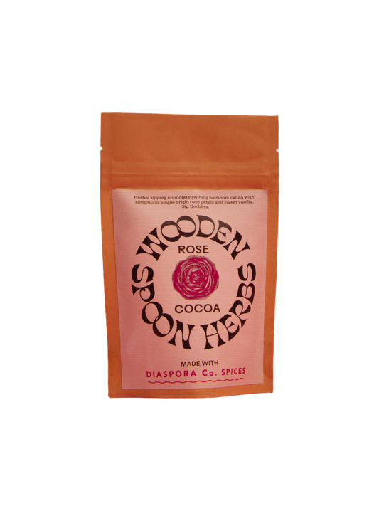 A tan packet with a pink label featuring the text "WOODEN SPOON HERBS Rose Cocoa, Made with Diaspora Co. Spices." A rose graphic is in the center of the label. This adaptogen-infused herbal spiced chocolate blends rose and heirloom cacao for a unique treat.