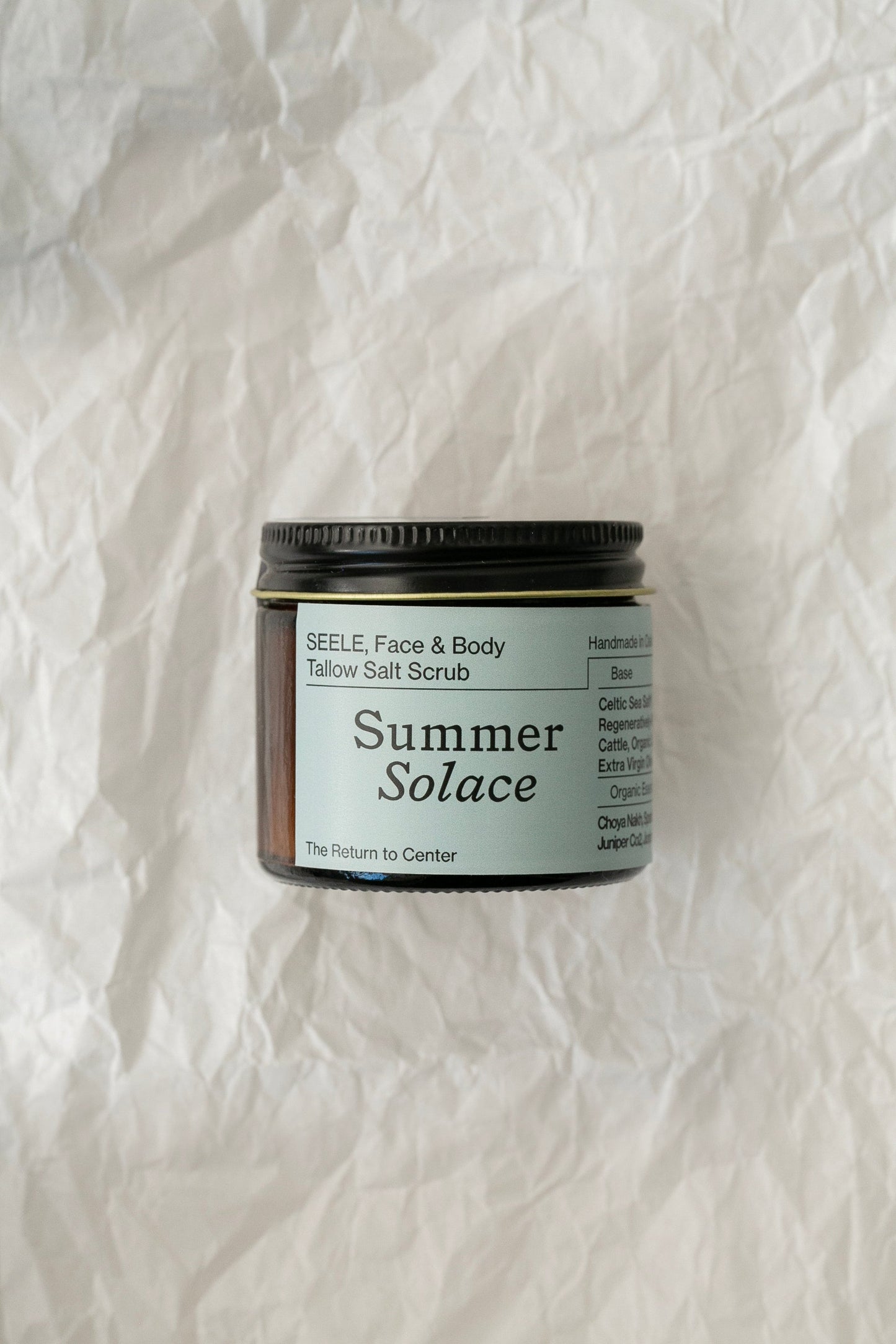 A jar of SEELE Celtic Sea Salt® Scrub - Regenerative Tallow® by Summer Solace Tallow sits on crumpled, textured white paper. The label highlights ingredients such as Celtic Sea Salt and coconut oil, infused with a hint of marine scent to invigorate your senses.