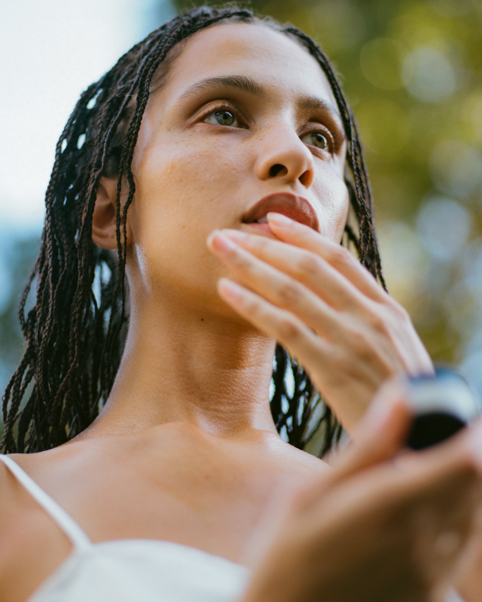 A person with braided hair looks off into the distance while touching their face, perhaps feeling the soothing effects of Surya's Calming Lip Therapy. They are holding the small tube of this product in their other hand. The background is blurred with green and blue hues, suggesting an outdoor setting with foliage.