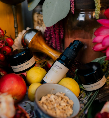 A vibrant arrangement of Surya skincare products, including a pump bottle labeled "balancing hydrating oil," and two round containers labeled "calming lip therapy" and "balancing collagen cream," surrounded by lemons, flowers, and various fruits in a lush, colorful setting. 