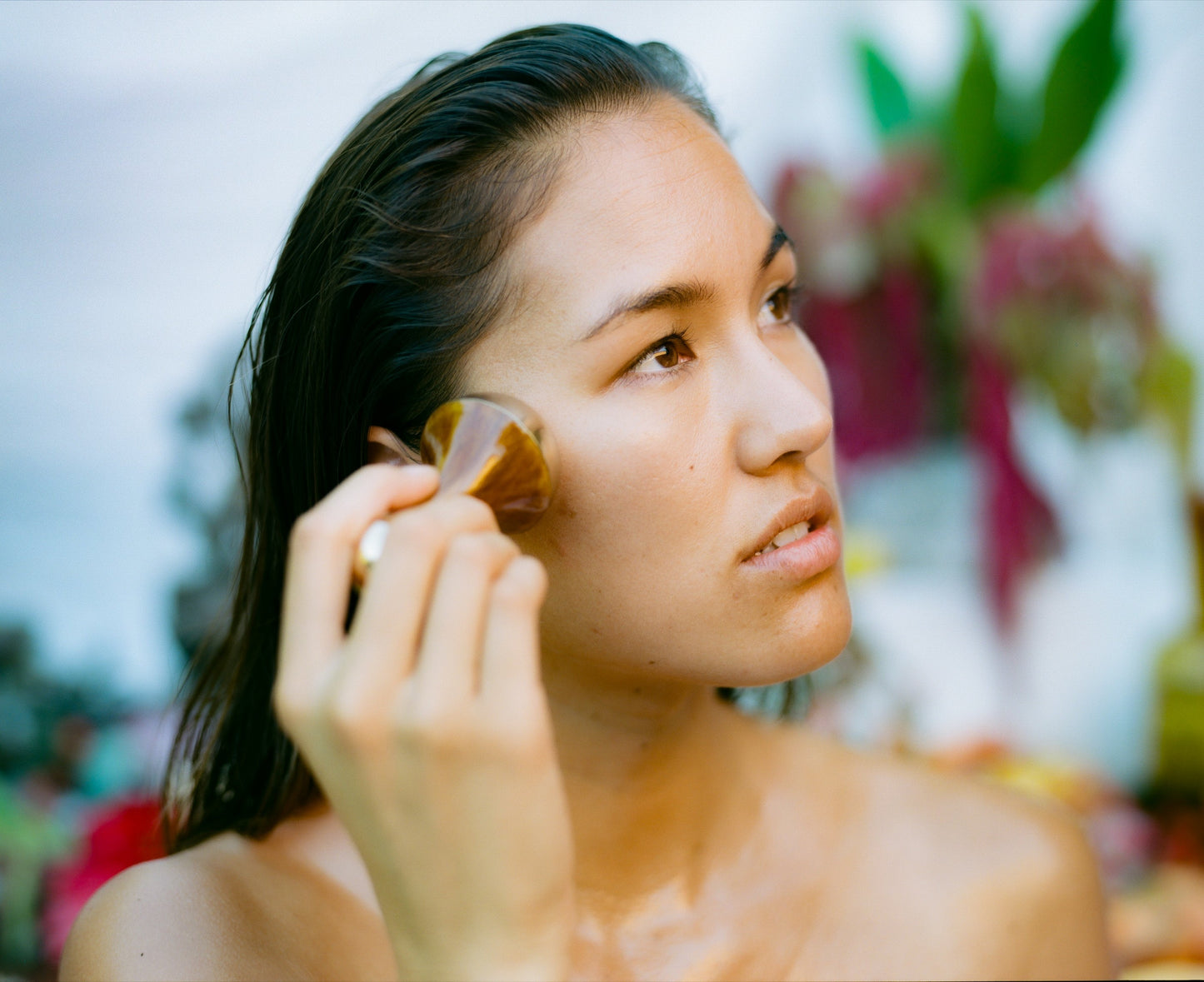 A person with long, dark wet hair applies makeup with a brush to their cheek. The background, blurred but vibrant, features lush tropical plants and flowers. Focused in the scene, they hold the brush in their right hand and a product from Surya's Skincare Discovery Set in the other, showcasing an Ayurvedic beauty routine amidst nature's serene backdrop.