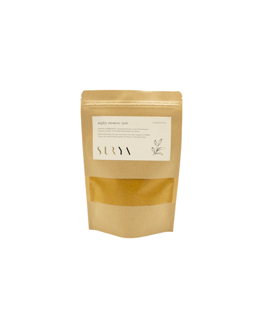 A brown kraft paper pouch labeled "Mighty Turmeric Latte" from Surya highlights its role in your wellness routine. Featuring white text, a simple plant design, and a transparent window showing the contents inside, this Ayurvedic latte mix prominently displays the brand name "Surya" on the label.