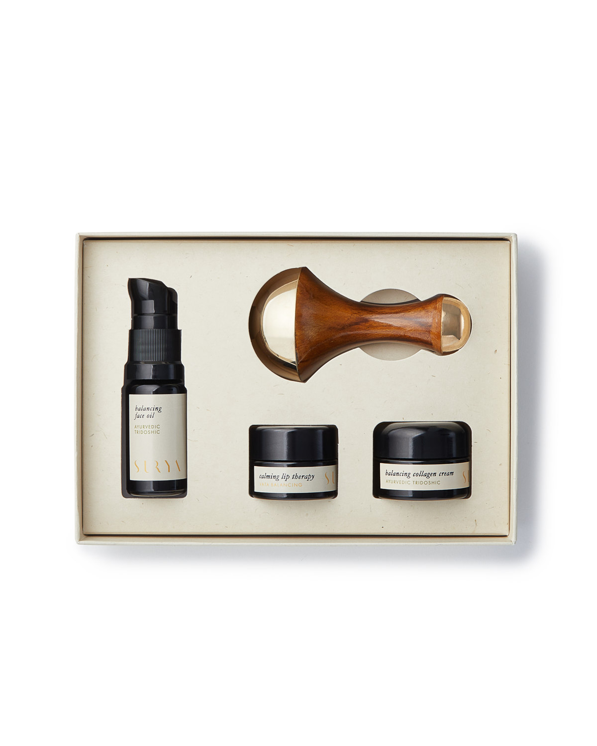 The Surya Skincare Discovery Set comes in a beige box and includes four items: a pump bottle labeled "balancing serum," two small jars labeled "lifting eye therapy" and "balancing collagen cream," and a wooden-handled Kansa Wand with a gold-colored roller head. This set provides an unparalleled Ayurvedic beauty experience.