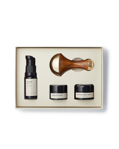 The Surya Skincare Discovery Set comes in a beige box and includes four items: a pump bottle labeled "balancing serum," two small jars labeled "lifting eye therapy" and "balancing collagen cream," and a wooden-handled Kansa Wand with a gold-colored roller head. This set provides an unparalleled Ayurvedic beauty experience.