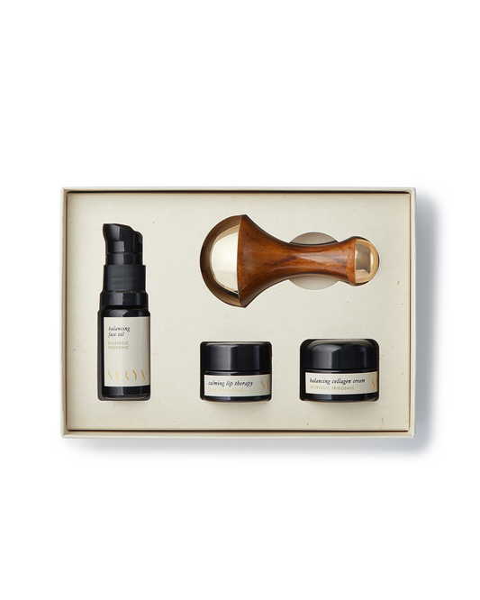 The Surya Skincare Discovery Set comes in a beige box and includes four items: a pump bottle labeled "balancing serum," two small jars labeled "lifting eye therapy" and "balancing collagen cream," and a wooden-handled Kansa Wand with a gold-colored roller head. This set provides an unparalleled Ayurvedic beauty experience.