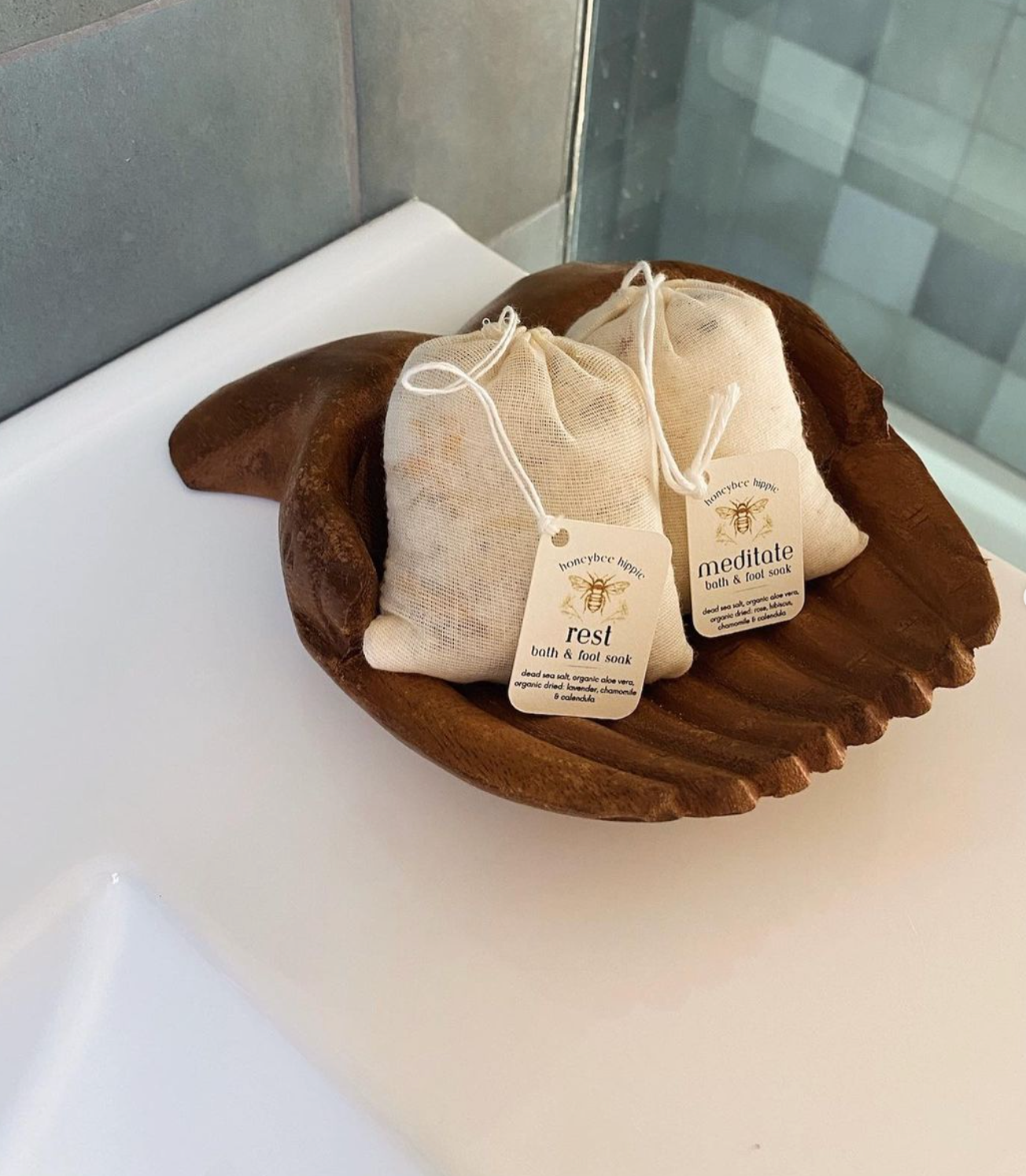 Two small burlap bags labeled “rest” and “meditate” sit on a wooden, leaf-shaped dish on a white surface. These bags contain bath soaks packed with organic herbs, perfect for relaxation. "Rest" is from Honeybee Hippie's line and makes an ideal addition to any unwinding routine.