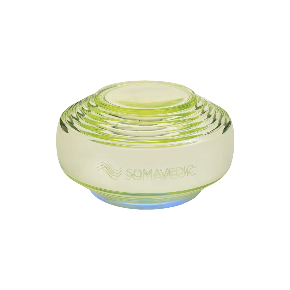 A translucent, greenish-yellow, disc-shaped device with layered ridges. The word "Somavedic USA Vedic" and a wave-like symbol are etched on the front, highlighting its role in water structuring. The base emits a soft blue light.