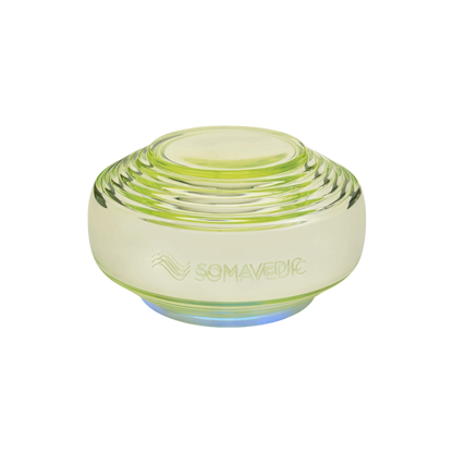 A translucent, greenish-yellow, disc-shaped device with layered ridges. The word "Somavedic USA Vedic" and a wave-like symbol are etched on the front, highlighting its role in water structuring. The base emits a soft blue light.