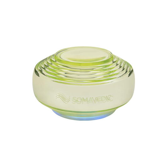 A translucent, greenish-yellow, disc-shaped device with layered ridges. The word "Somavedic USA Vedic" and a wave-like symbol are etched on the front, highlighting its role in water structuring. The base emits a soft blue light.