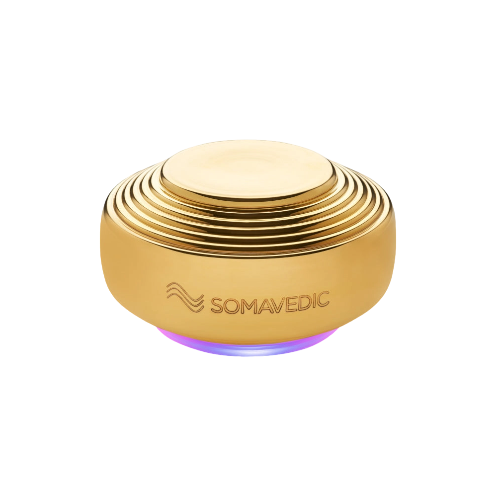 A round, gold-colored device with a ribbed design, and "Somavedic USA" engraved on the side. The Somavedic Gold uses quantum physics technology to emit a soft purple glow from its base, offering EMF radiation protection.