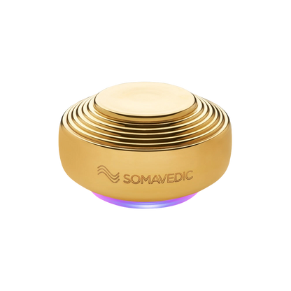 A round, gold-colored device with a ribbed design, and "Somavedic USA" engraved on the side. The Somavedic Gold uses quantum physics technology to emit a soft purple glow from its base, offering EMF radiation protection.