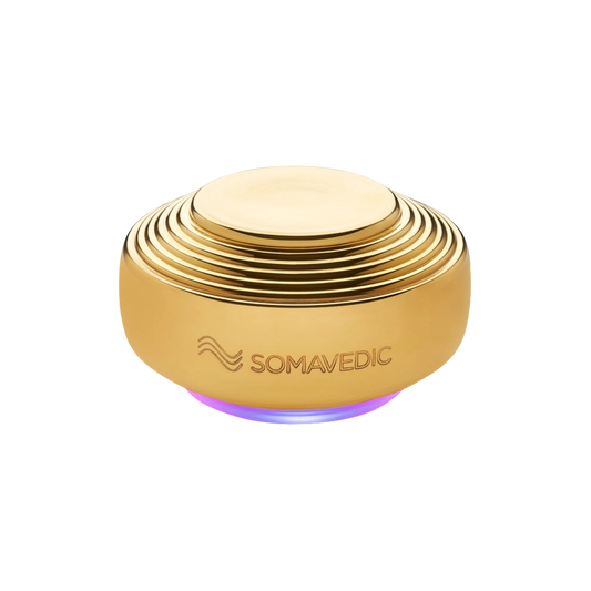 A round, gold-colored device with a ribbed design, and "Somavedic USA" engraved on the side. The Somavedic Gold uses quantum physics technology to emit a soft purple glow from its base, offering EMF radiation protection.