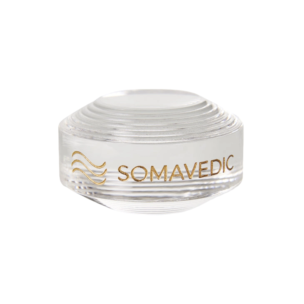 A clear, round glass device named "Tiny" by Somavedic USA is displayed on a white background with the word "SOMAVEDIC" and a stylized wave design printed in gold on the side. The top features a series of concentric circular ridges, creating a tiny yet charged protective field.