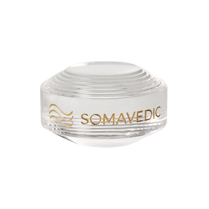 A clear, round glass device named "Tiny" by Somavedic USA is displayed on a white background with the word "SOMAVEDIC" and a stylized wave design printed in gold on the side. The top features a series of concentric circular ridges, creating a tiny yet charged protective field.