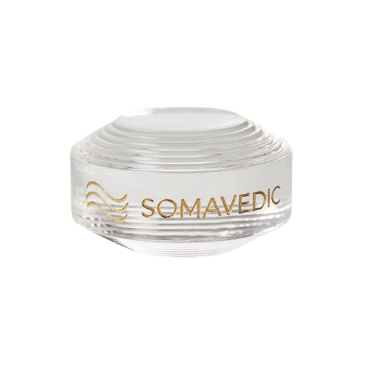 A clear, round glass device named "Tiny" by Somavedic USA is displayed on a white background with the word "SOMAVEDIC" and a stylized wave design printed in gold on the side. The top features a series of concentric circular ridges, creating a tiny yet charged protective field.