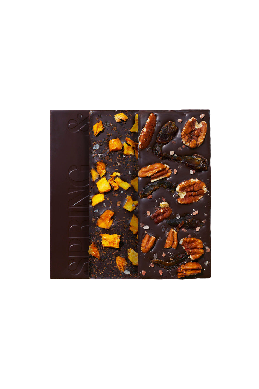 A bar of dark chocolate from the Spice Market Collection by Spring & Mulberry is divided into three sections. The left section is plain, embossed with the word "SPRING," the middle is topped with naturally sweet dried mango pieces, and the right section has a variety of nuts and seeds.